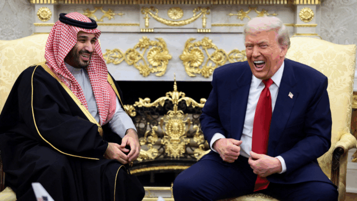 Dois homens sentados em poltronas claras conversam e riem em uma sala ornamentada com detalhes dourados; um deles veste terno azul com gravata vermelha, e o outro usa túnica tradicional preta com detalhes dourados e um lenço estampado vermelho e branco na cabeça.