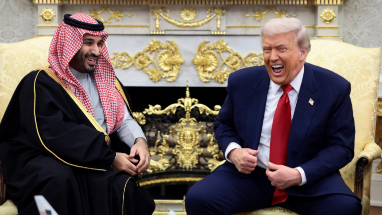 Dois homens sentados em poltronas claras conversam e riem em uma sala ornamentada com detalhes dourados; um deles veste terno azul com gravata vermelha, e o outro usa túnica tradicional preta com detalhes dourados e um lenço estampado vermelho e branco na cabeça.