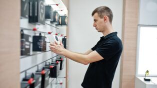 Homem branco e loiro de camiseta preta analisando produtos a venda em uma loja de eletrônicos