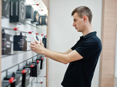 Homem branco e loiro de camiseta preta analisando produtos a venda em uma loja de eletrônicos