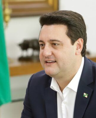 Ratinho Junior sentado em seu gabinete, com bandeira do Paraná ao fundo