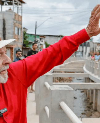 Homem de camisa vermelha e chapéu de palha acena sorridente em uma rua com obras e casas simples ao fundo, enquanto várias pessoas observam e registram o momento.