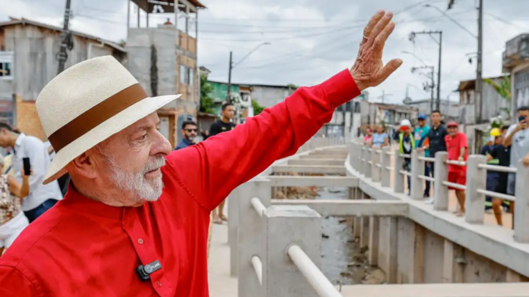 Homem de camisa vermelha e chapéu de palha acena sorridente em uma rua com obras e casas simples ao fundo, enquanto várias pessoas observam e registram o momento.