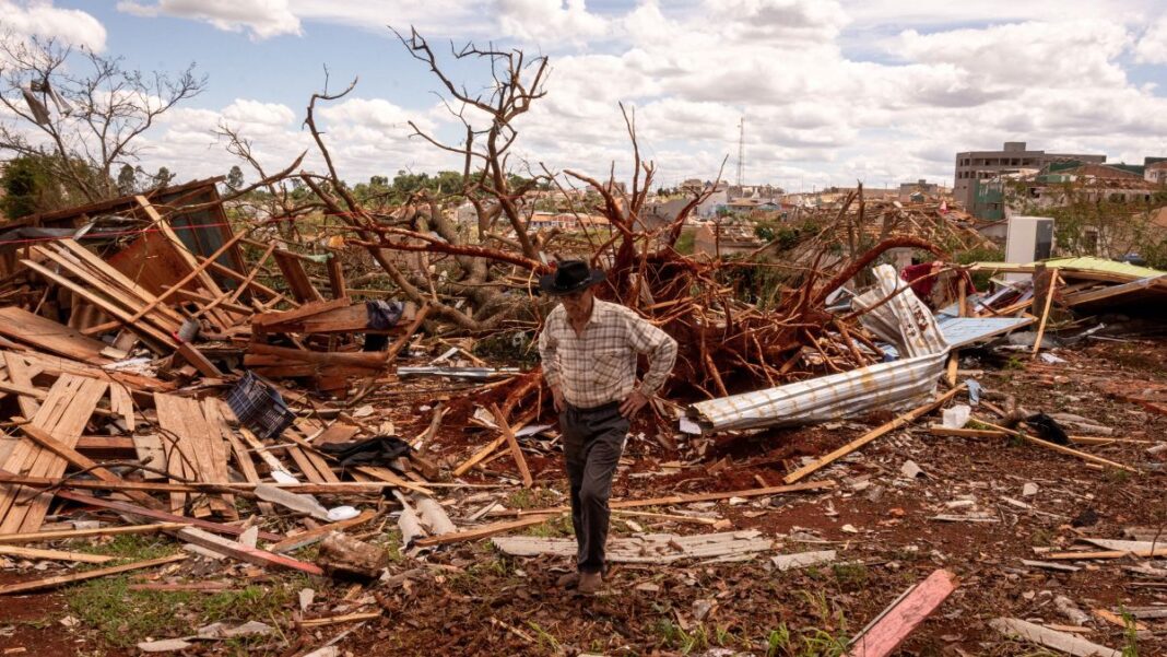 Homem de chapéu caminha entre escombros após a destruição causada por um evento climático severo. Ao fundo, árvores arrancadas e casas destruídas mostram a extensão dos danos sob um céu parcialmente nublado.
