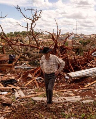 Homem de chapéu caminha entre escombros após a destruição causada por um evento climático severo. Ao fundo, árvores arrancadas e casas destruídas mostram a extensão dos danos sob um céu parcialmente nublado.