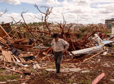 Homem de chapéu caminha entre escombros após a destruição causada por um evento climático severo. Ao fundo, árvores arrancadas e casas destruídas mostram a extensão dos danos sob um céu parcialmente nublado.