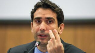 Presidente do Banco Central, Gabriel Galípolo fala ao microfone e faz sinal de número dois com os dedos das mãos esquerda