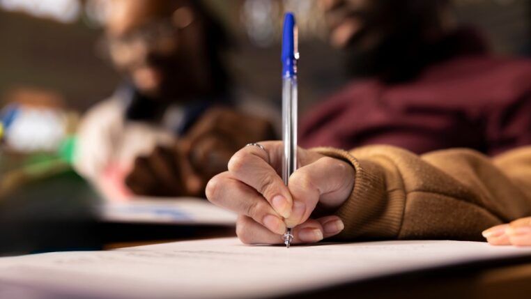 Mão de mulher negra segurando uma caneta azul e redigindo um texto