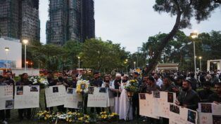 Dezenas de chineses carregam cartazes e flores com o prédio incendiado ao fundo