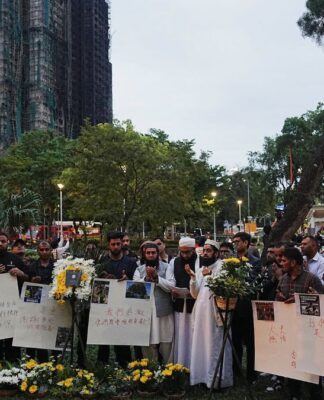 Dezenas de chineses carregam cartazes e flores com o prédio incendiado ao fundo