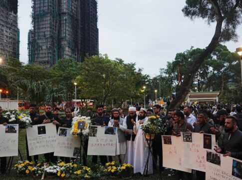 Dezenas de chineses carregam cartazes e flores com o prédio incendiado ao fundo