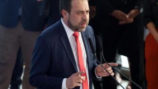 Boulos discursando em um palanque usa ndo terno escuro, camisa branca e gravata vermelha