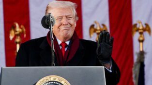 Donald Trump sorri diante de microfone com a bandeira dos EUA ao fundo