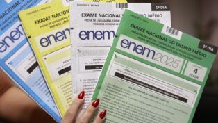 Mão segurando ps quatro cadernos de perguntas do ENEM. Um azul, um amarelo, um branco e um verde.