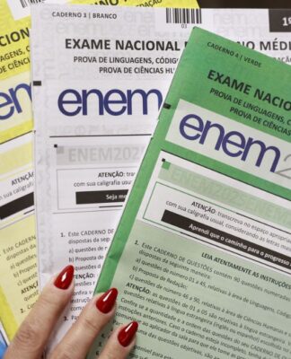 Mão segurando ps quatro cadernos de perguntas do ENEM. Um azul, um amarelo, um branco e um verde.