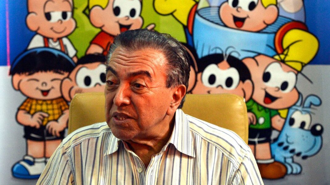 Maurício de Sousa sentado à frente de uma mesa, com seus personagens desenhados ao fundo