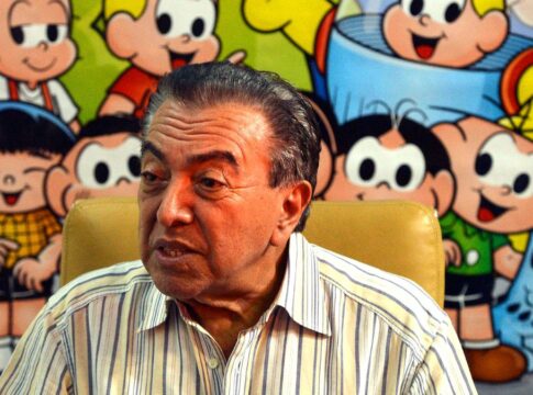 Maurício de Sousa sentado à frente de uma mesa, com seus personagens desenhados ao fundo