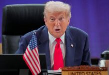 Trump em sua mesa na Casa Branca, falando. Ele usa gravata vemelha, terno azul e camisa branca.