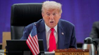 Trump em sua mesa na Casa Branca, falando. Ele usa gravata vemelha, terno azul e camisa branca.