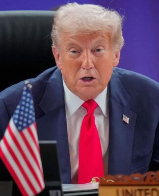 Trump em sua mesa na Casa Branca, falando. Ele usa gravata vemelha, terno azul e camisa branca.