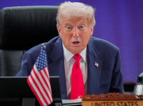 Trump em sua mesa na Casa Branca, falando. Ele usa gravata vemelha, terno azul e camisa branca.