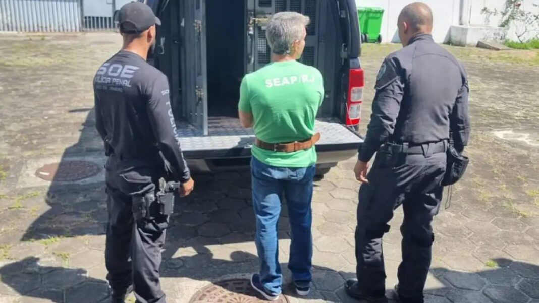 Rogério Andrade está de costas, de camiseta verde, diante de um camburão da polícia