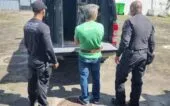 Rogério Andrade está de costas, de camiseta verde, diante de um camburão da polícia