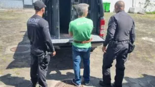 Rogério Andrade está de costas, de camiseta verde, diante de um camburão da polícia