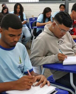 Foto de uma sala de aula. No plano frontal, um aluno negro e um aluno pardo fazendo anotações em seus cadernos