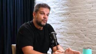 Rodrigo Pimentel sentado em uma mesa, dando entrevitsa. Ele veste camiseta preta e está ao lado de uma parede de tijolinho branco