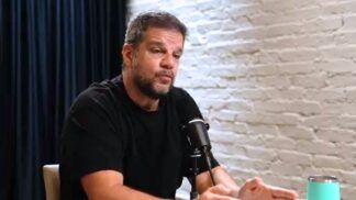 Rodrigo Pimentel sentado em uma mesa, dando entrevitsa. Ele veste camiseta preta e está ao lado de uma parede de tijolinho branco