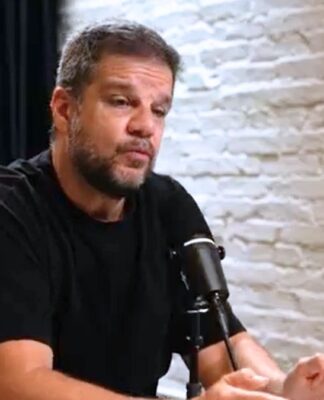 Rodrigo Pimentel sentado em uma mesa, dando entrevitsa. Ele veste camiseta preta e está ao lado de uma parede de tijolinho branco