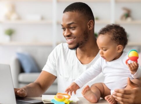 Homem negro sentando de frente para um notebook, com um bebê ao lado