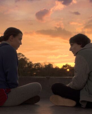 Garota e garoto sentados, de pernas cruzadas e de lado, trocando olhares sob o pôr-do-sol