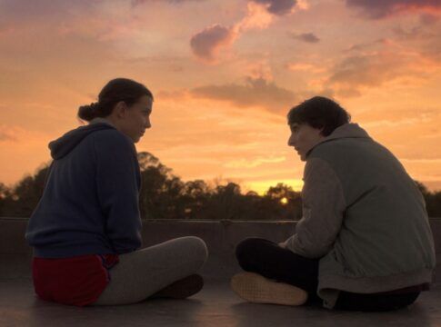 Garota e garoto sentados, de pernas cruzadas e de lado, trocando olhares sob o pôr-do-sol