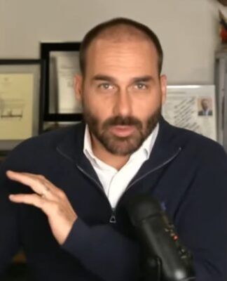 Eduardo Bolsonaro participando remotamente de uma sessão plenária. Ele está sentado à mesa com um microfone cardioide ao seu lado