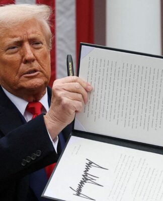 Donald Trump segura uma caneta com a mão direita e ergue texto de lei que assinou