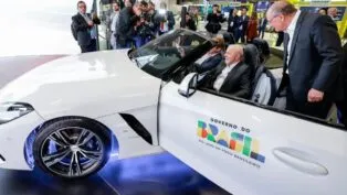 Lula no salão do automóvel, dentro de um carro branco, com a porta aberta com logotipo do governo federal. ao lado, Geraldo Alckimn observa o interior do veículo.