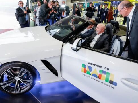 Lula no salão do automóvel, dentro de um carro branco, com a porta aberta com logotipo do governo federal. ao lado, Geraldo Alckimn observa o interior do veículo.