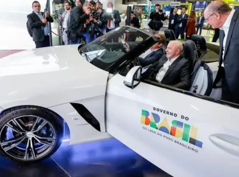Lula no salão do automóvel, dentro de um carro branco, com a porta aberta com logotipo do governo federal. ao lado, Geraldo Alckimn observa o interior do veículo.