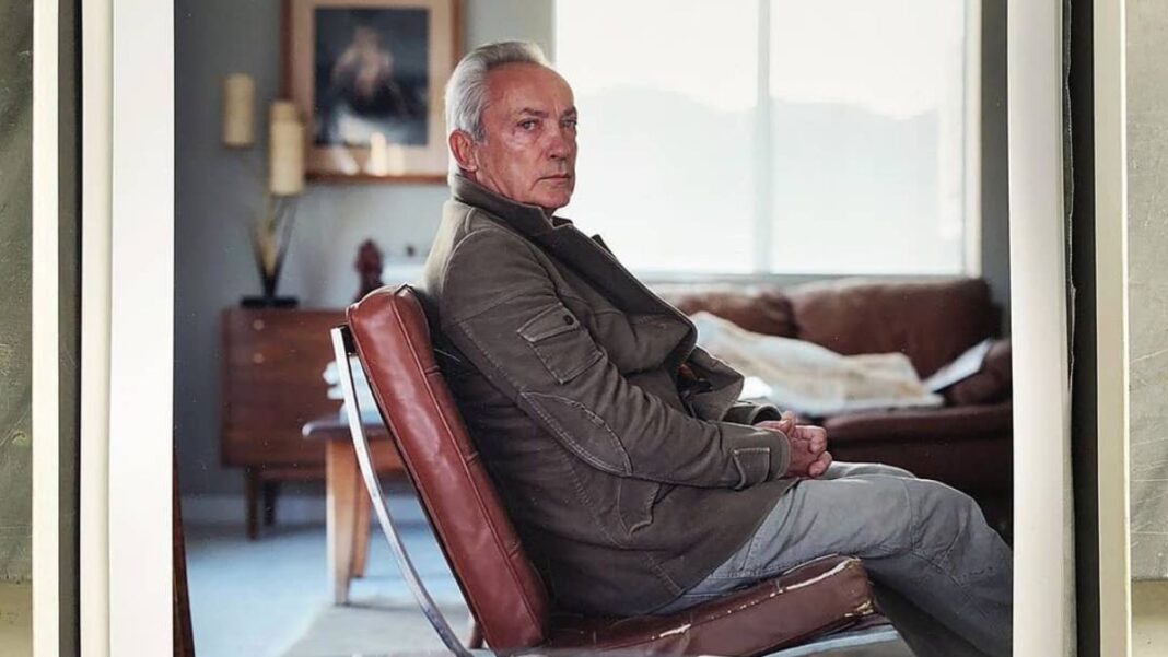 Ator Udo Kier veste um casaco e está sentado numa poltrona olhando de lado