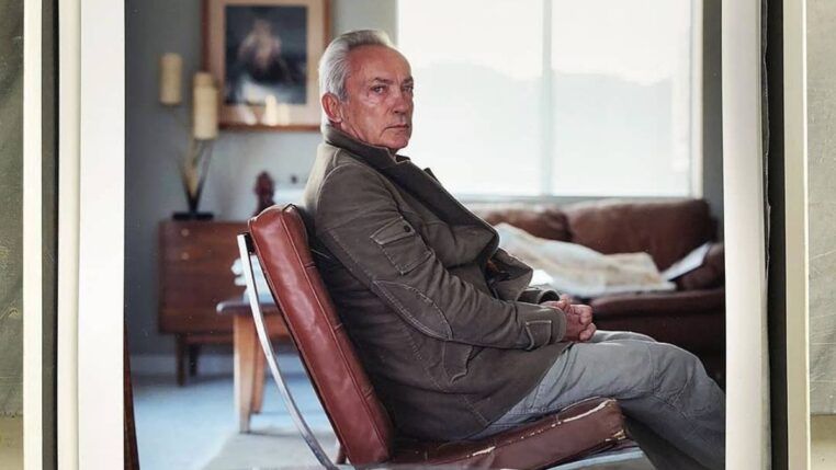 Ator Udo Kier veste um casaco e está sentado numa poltrona olhando de lado