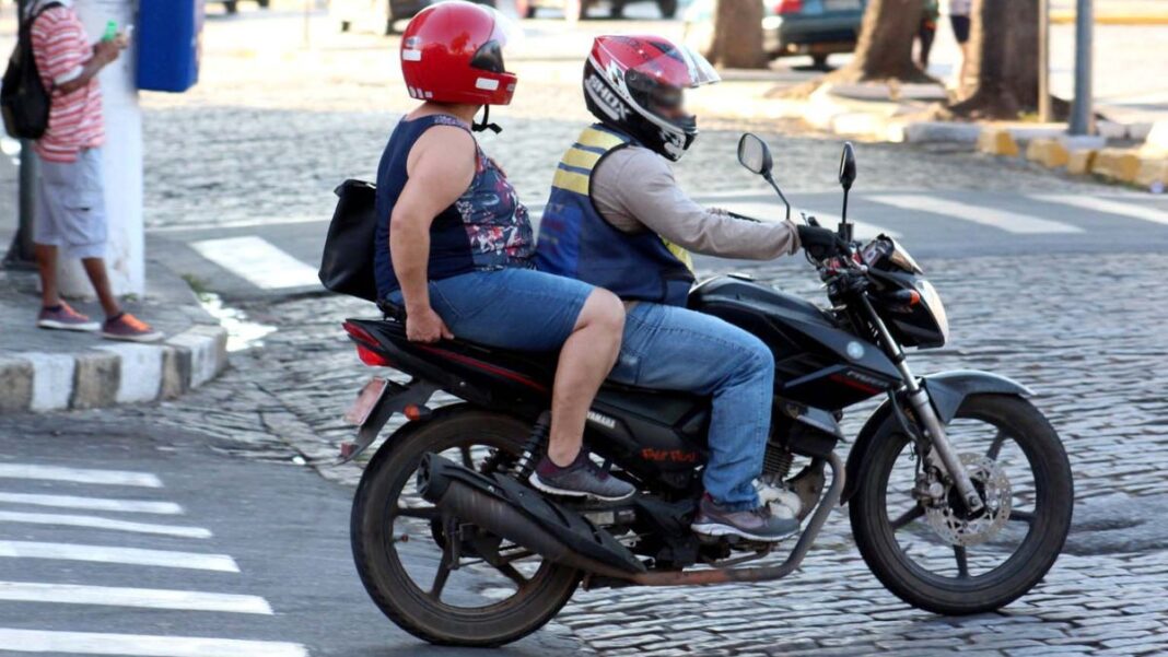 Duas pessoas estão em uma moto circulando por uma rua de paralelepípedos. O condutor veste capacete vermelho com viseira e colete refletivo, enquanto a passageira usa camiseta estampada, capacete vermelho e mochila nas costas. Ambos estão de jeans. Ao fundo, há um pedestre usando celular e a calçada com pintura preta e branca.