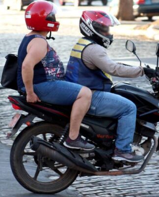 Duas pessoas estão em uma moto circulando por uma rua de paralelepípedos. O condutor veste capacete vermelho com viseira e colete refletivo, enquanto a passageira usa camiseta estampada, capacete vermelho e mochila nas costas. Ambos estão de jeans. Ao fundo, há um pedestre usando celular e a calçada com pintura preta e branca.