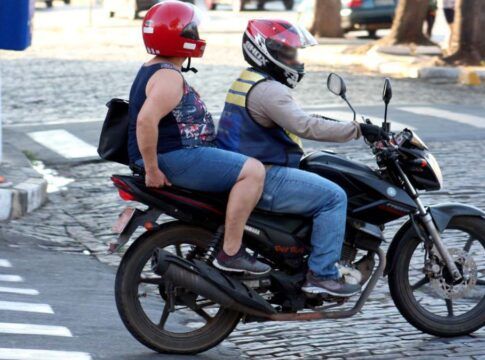 Duas pessoas estão em uma moto circulando por uma rua de paralelepípedos. O condutor veste capacete vermelho com viseira e colete refletivo, enquanto a passageira usa camiseta estampada, capacete vermelho e mochila nas costas. Ambos estão de jeans. Ao fundo, há um pedestre usando celular e a calçada com pintura preta e branca.