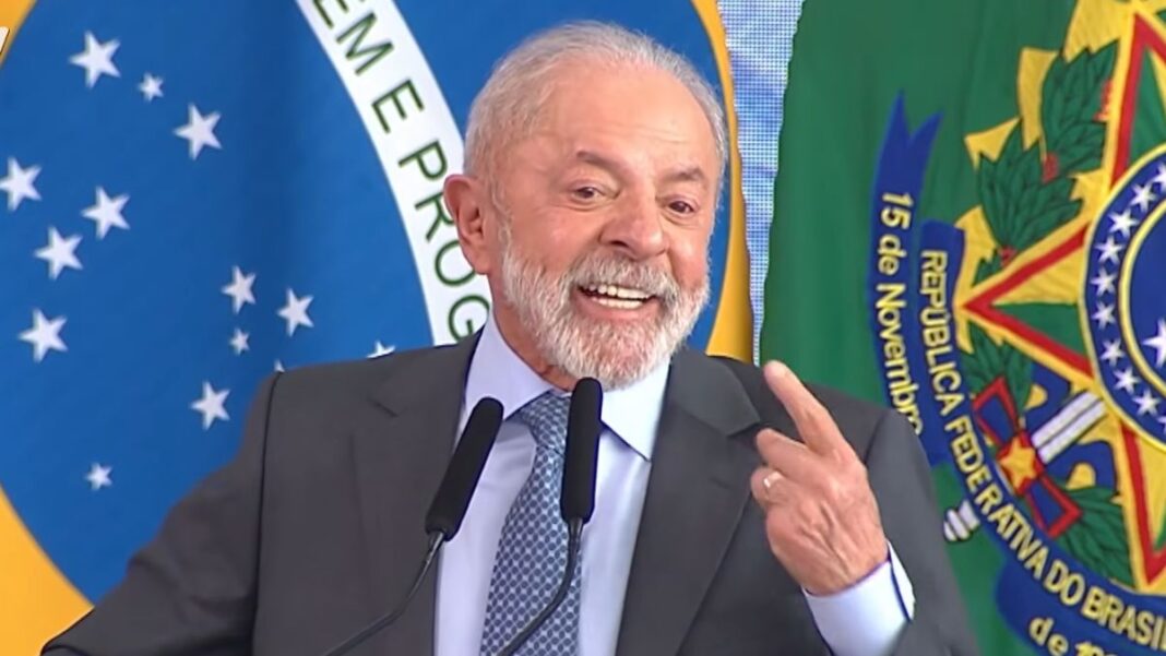 Presidente Lula, durante seu discurso, sorrindo e apontando com o dedo indicador para si mesmo. Ao fundo estão a bandeira do Brasil e cortinas azuis