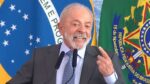 Presidente Lula, durante seu discurso, sorrindo e apontando com o dedo indicador para si mesmo. Ao fundo estão a bandeira do Brasil e cortinas azuis