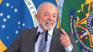 Presidente Lula, durante seu discurso, sorrindo e apontando com o dedo indicador para si mesmo. Ao fundo estão a bandeira do Brasil e cortinas azuis