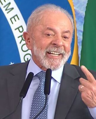 Presidente Lula, durante seu discurso, sorrindo e apontando com o dedo indicador para si mesmo. Ao fundo estão a bandeira do Brasil e cortinas azuis