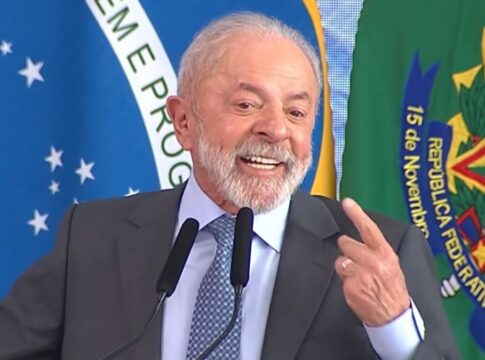 Presidente Lula, durante seu discurso, sorrindo e apontando com o dedo indicador para si mesmo. Ao fundo estão a bandeira do Brasil e cortinas azuis